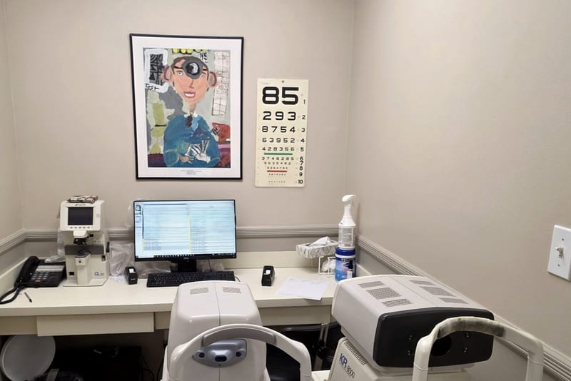 The pre-testing room, with an autorefractor and a lensmeter on the counter beside a computer workstation, and a wall-mounted eye chart.
